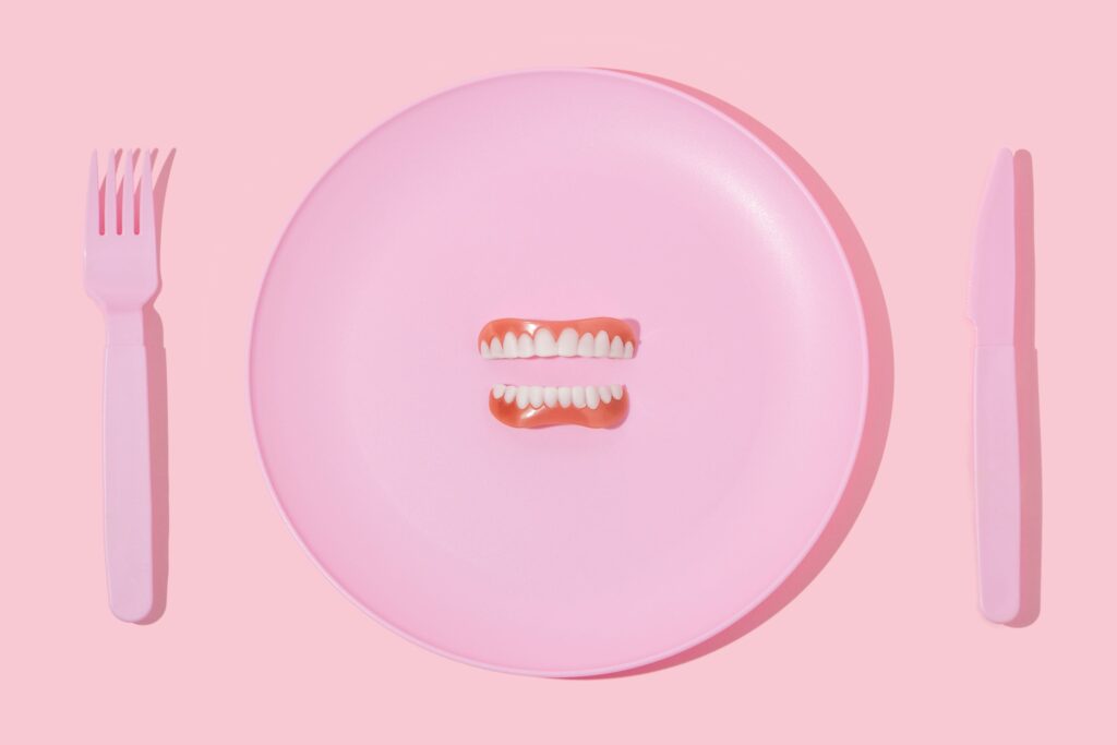 Full set of dentures on a pink plate between pink utensils on a lighter pink surface